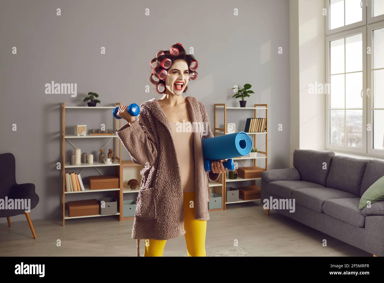 Glückliche Frau im Wohnzimmer stehend mit Matte und Hanteln Für Heimfitness-Workout Stockfoto
