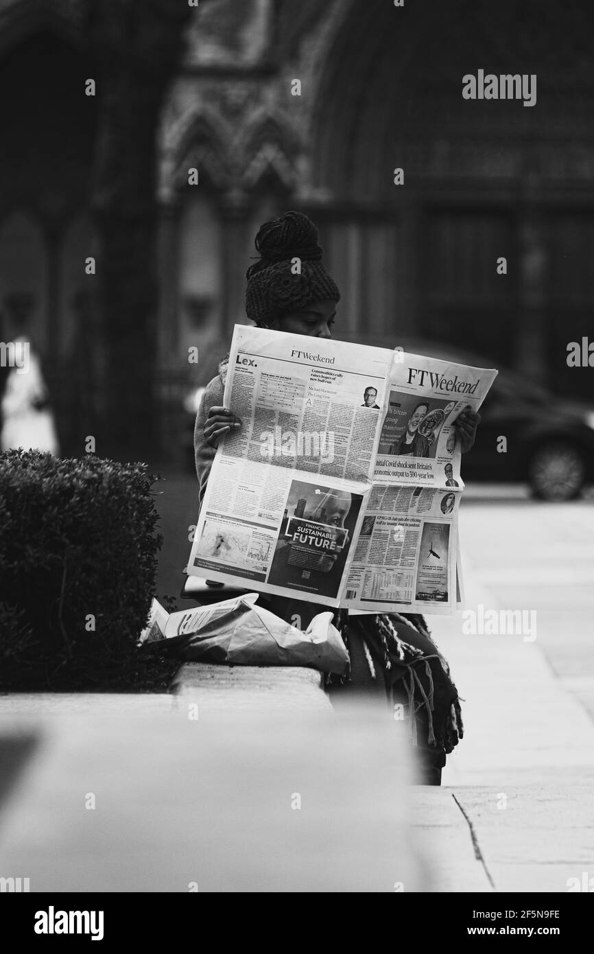 London UK Februar 2021 Vertikale Schwarz-Weiß-Aufnahme einer jungen Frau, die in einem öffentlichen Park sitzt, Zeitungen hält und liest Stockfoto