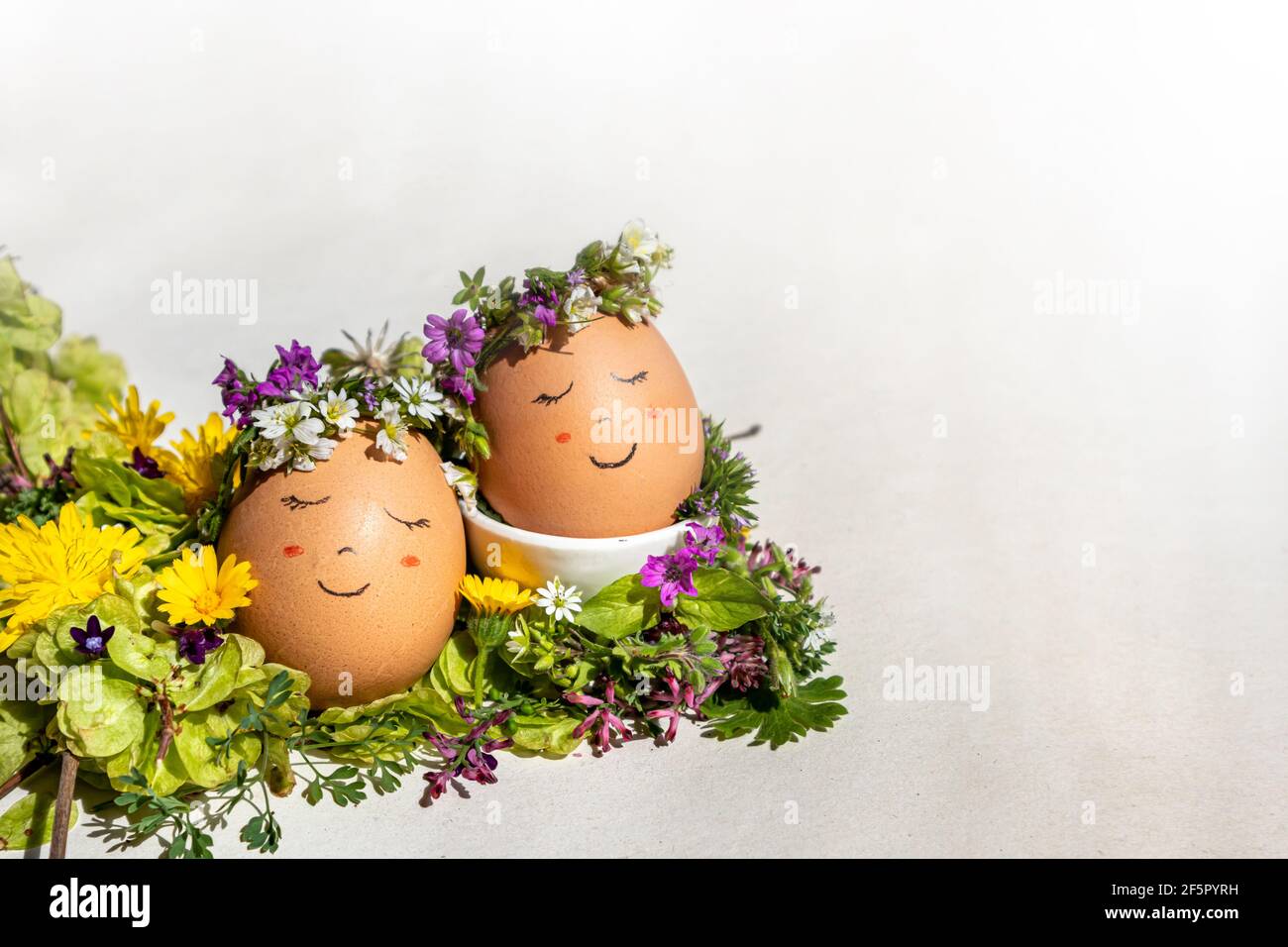 Ostern und Frühling Konzept. Zwei Eier mit einem lächelnden Gesicht und einer Blumengirlande, die die Frühlingssonne auf einem Bett aus Ulmenkernen und Wildblumen genießen Stockfoto