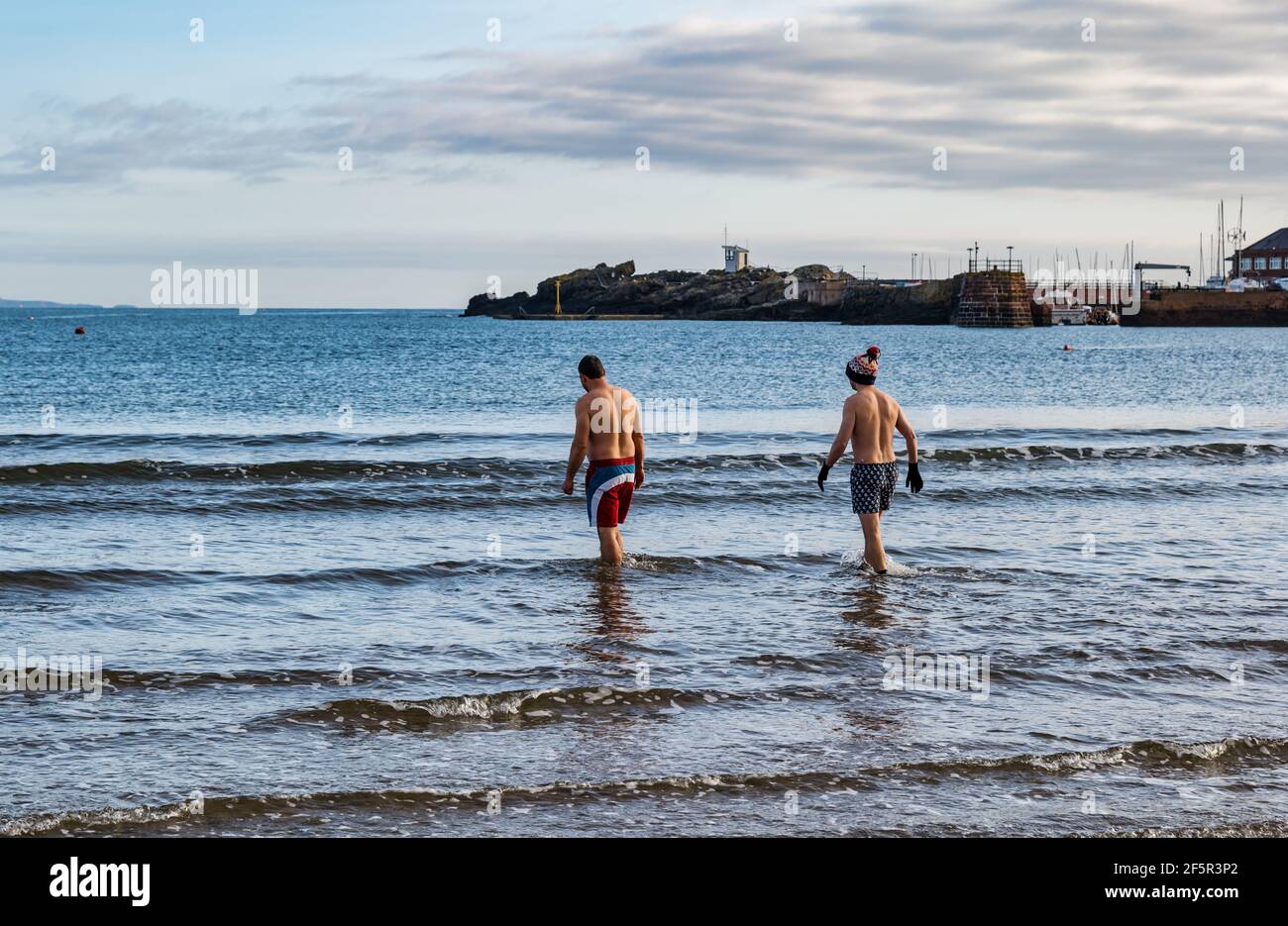 Zwei männliche Wildwasser- oder Freiwasserschwimmer mit Badehosen betreten die Firth of Forth Sea in West Bay, North Berwick, East Lothian, Schottland, Großbritannien Stockfoto