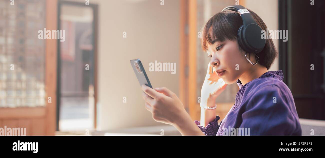 Glückliche junge asiatische Frau, die Musik mit Kopfhörern über Smartphone hört und Spaß hat, während sie an der Straße sitzt. Hintergrund des Banners Stockfoto