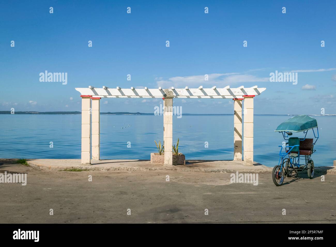 An einem sonnigen Tag in Punta Gorda, Kuba, sitzt ein blaues Dreirad-Taxi neben einem weißen Gitter am Ufer der Bucht von Cienfuegos. Stockfoto