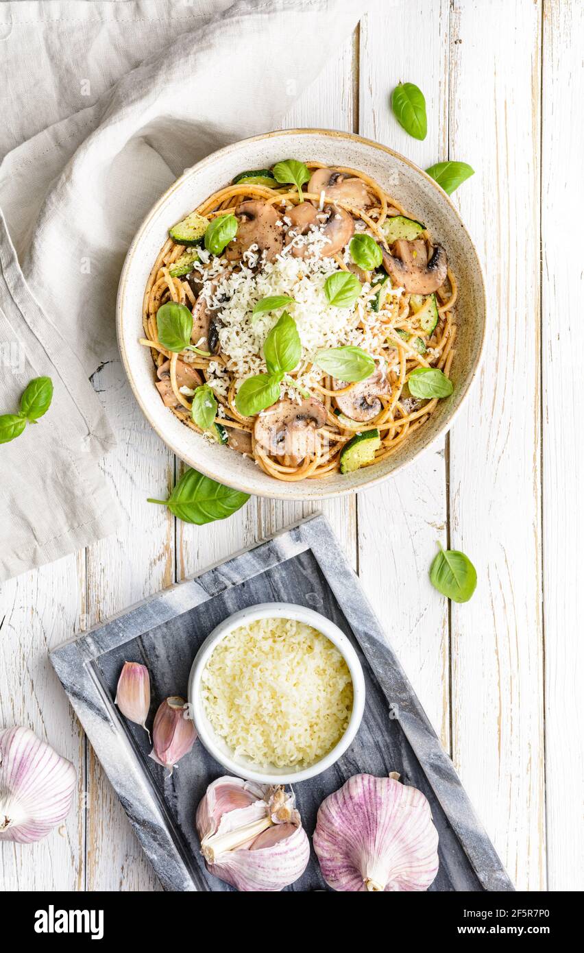 Gesunde Vollkornspaghetti mit sautierten Pilzen Knoblauch und Zucchini, gekrönt mit geriebenem Käse und Basilikumblättern auf rustikalem Holzhintergrund Stockfoto