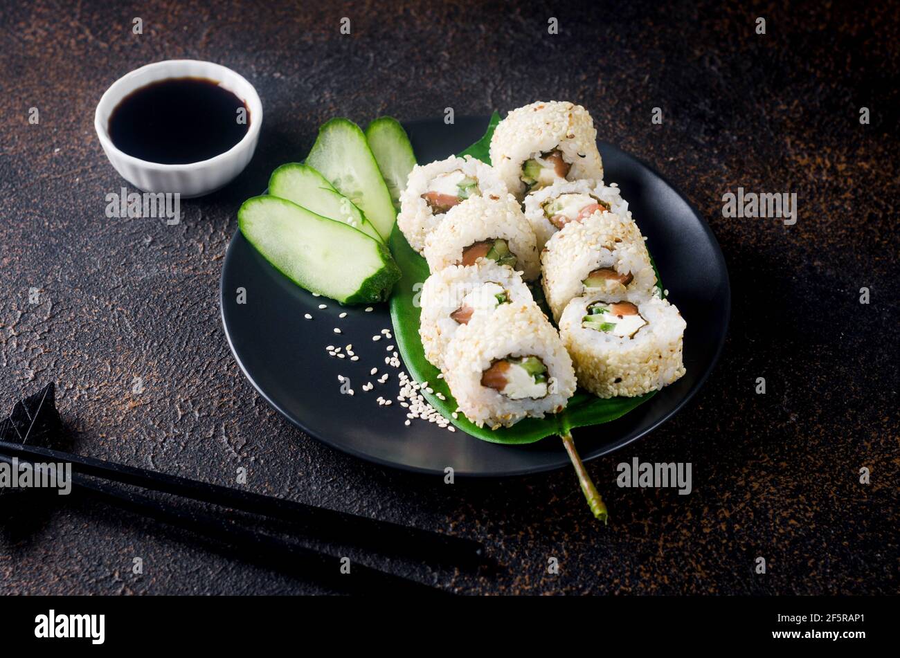 Leckeres Philadelphia Roll Sushi mit Lachs und Frischkäse in Sesam auf schwarzem Teller mit Sojasauce, Ingwer, Wasabi und Essstäbchen auf dunklem Tisch. Sushi Stockfoto