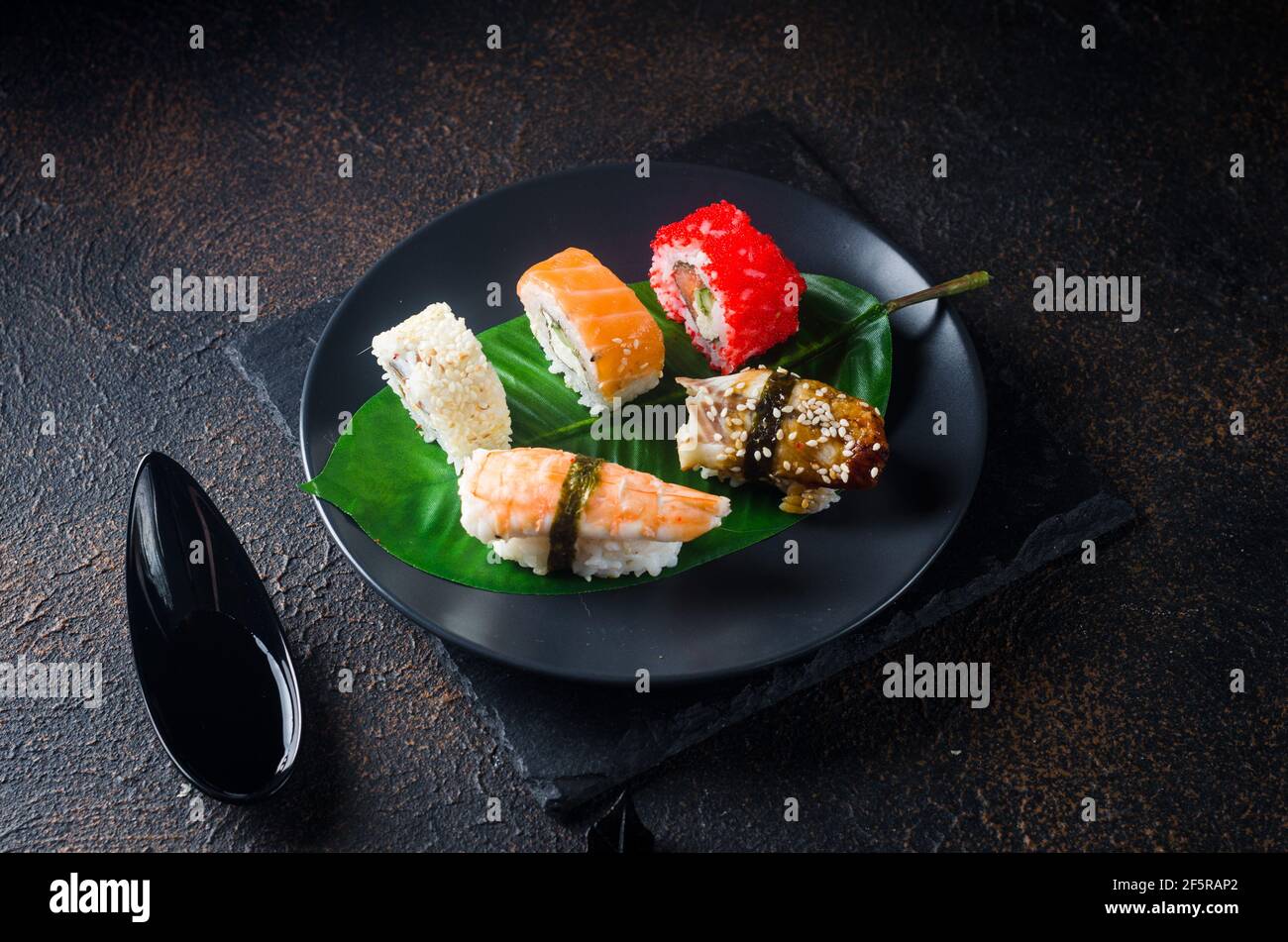 Leckere Sushi-Brötchen auf schwarzem Teller mit Saucen, Essstäbchen, Ingwer und Wasabi auf dunklem Tisch. Sushi-Menü. Lieferservice Japanisches Essen . Verschiedene Stockfoto