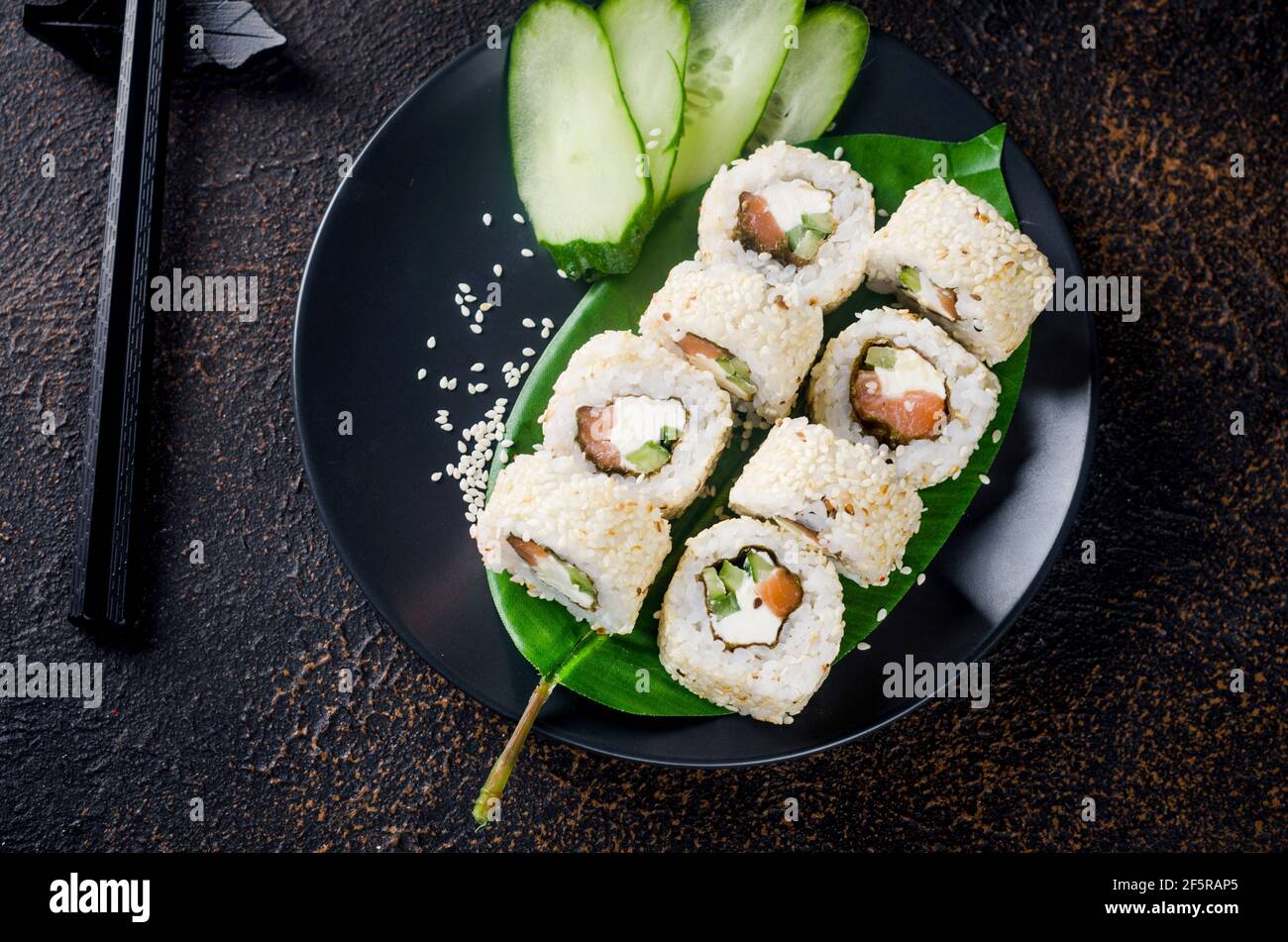 Leckeres Philadelphia Roll Sushi mit Lachs und Frischkäse in Sesam auf schwarzem Teller mit Sojasauce, Ingwer, Wasabi und Essstäbchen auf dunklem Tisch. Sushi Stockfoto