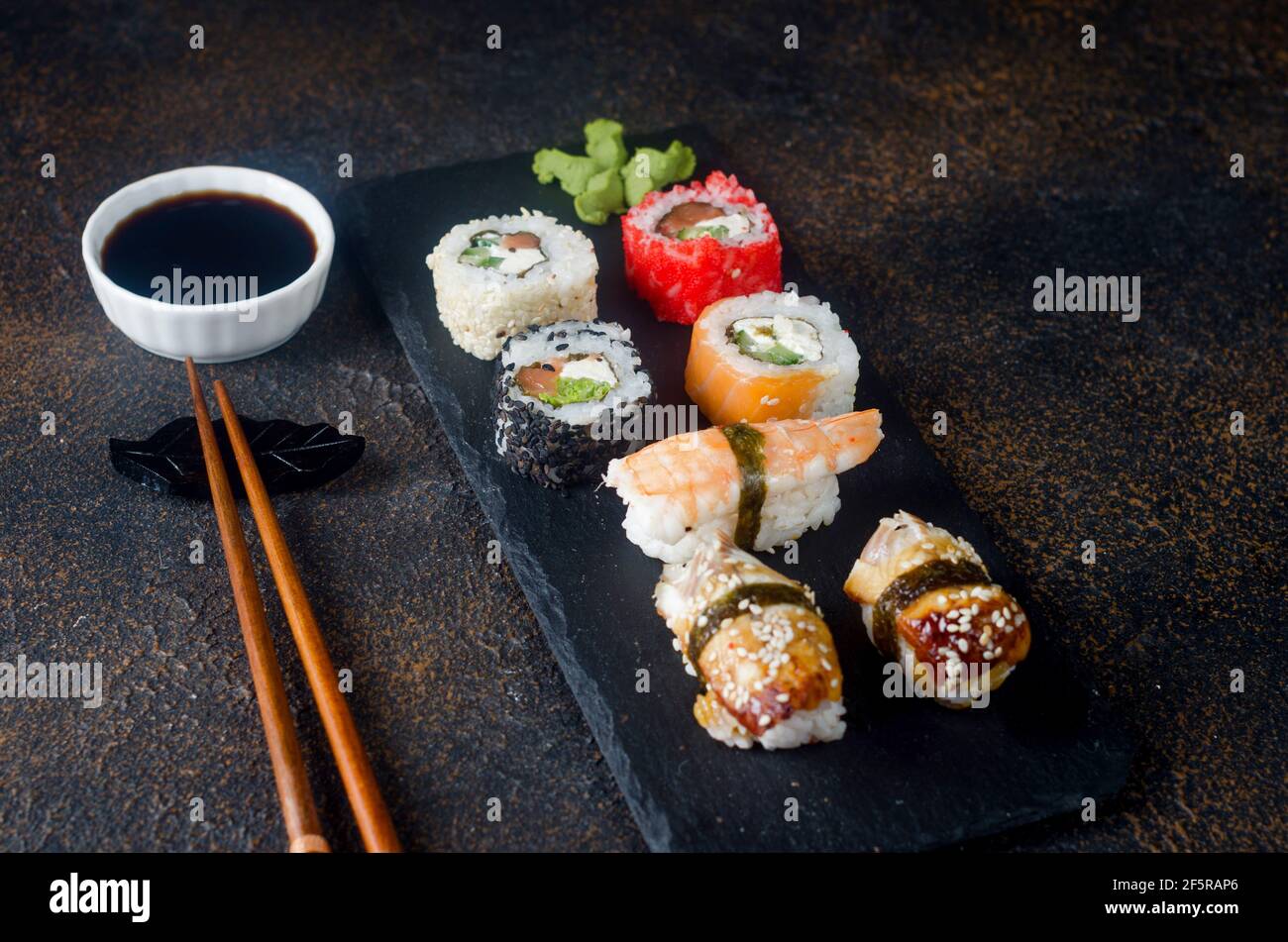 Leckere Sushi-Brötchen auf schwarzem Teller mit Saucen, Essstäbchen, Ingwer und Wasabi auf dunklem Tisch. Sushi-Menü. Lieferservice Japanisches Essen . Verschiedene Stockfoto