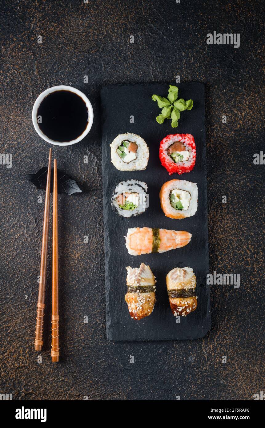 Leckere Sushi-Brötchen auf schwarzem Teller mit Saucen, Essstäbchen, Ingwer und Wasabi auf dunklem Tisch. Sushi-Menü. Lieferservice Japanisches Essen . Verschiedene Stockfoto
