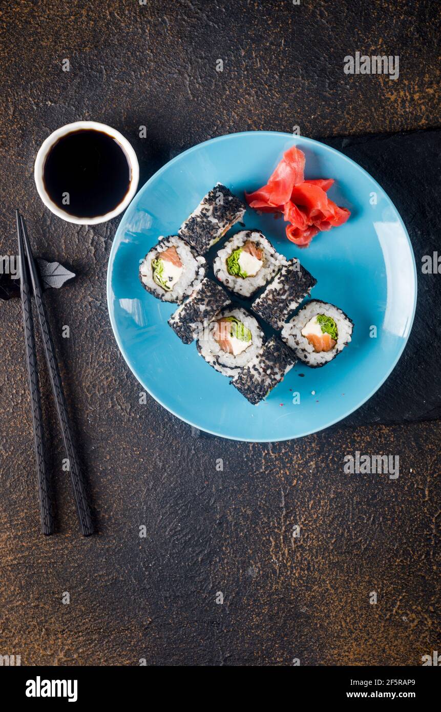 Leckere Sushi-Brötchen in schwarzem Sesam auf blauem Teller mit schwarzen Essstäbchen, Ingwer und Wasabi auf dunklem Hintergrund. Sushi-Menü. Lieferservice Japanisch Stockfoto