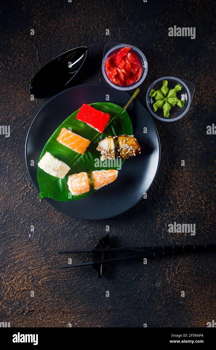 Leckere Sushi-Brötchen auf schwarzem Teller mit Saucen, Essstäbchen, Ingwer und Wasabi auf dunklem Tisch. Sushi-Menü. Lieferservice Japanisches Essen . Verschiedene Stockfoto