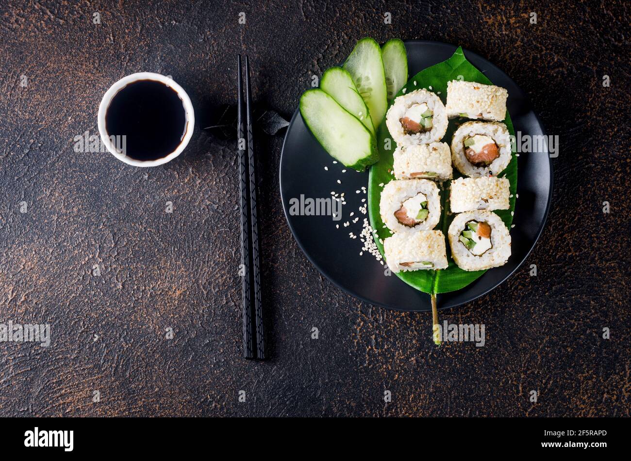 Leckeres Philadelphia Roll Sushi mit Lachs und Frischkäse in Sesam auf schwarzem Teller mit Sojasauce, Ingwer, Wasabi und Essstäbchen auf dunklem Tisch. Sushi Stockfoto