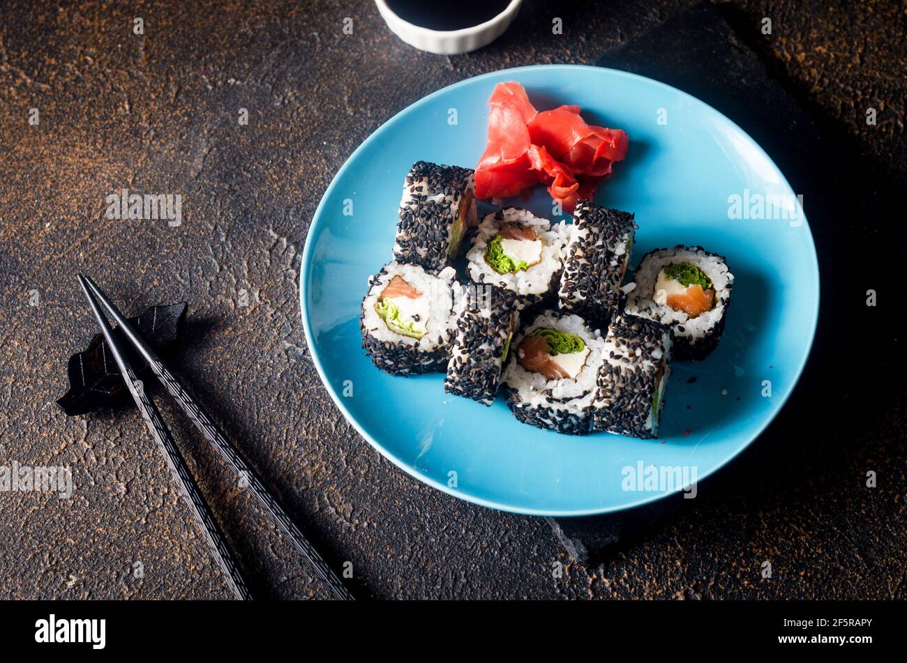 Leckere Sushi-Brötchen in schwarzem Sesam auf blauem Teller mit schwarzen Essstäbchen, Ingwer und Wasabi auf dunklem Hintergrund. Sushi-Menü. Lieferservice Japanisch Stockfoto