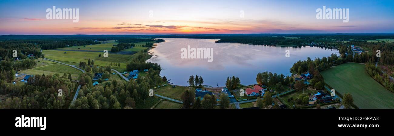 Luftlandschaftliches Panorama der Mitternachtssonne in Nordschweden, mittelsommerliche helle Nächte, Polarregion. Rot-orange Himmel, blauer See, schwedische Häuser bei coas Stockfoto