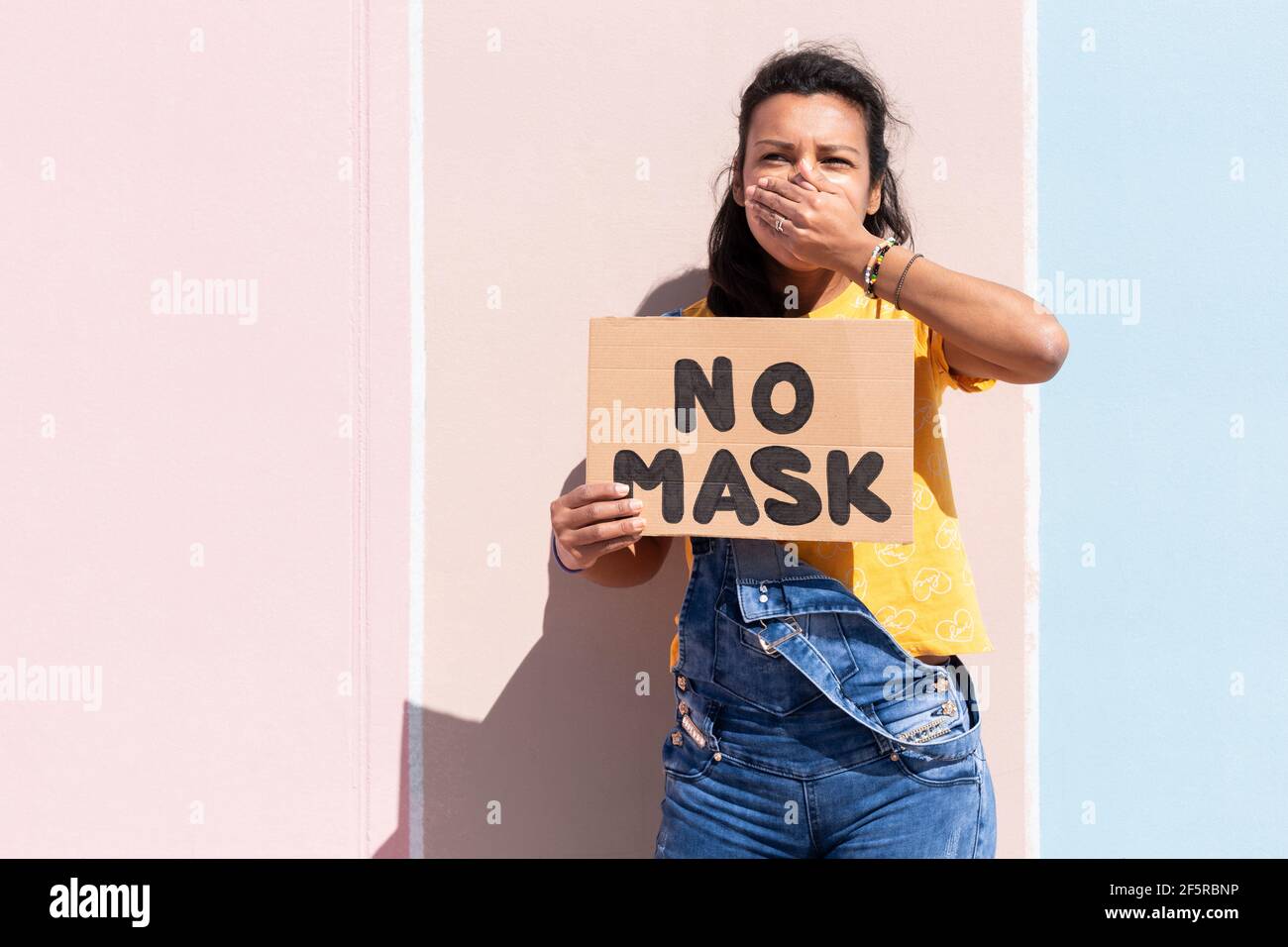 Porträt einer hispanischen Frau, die ein Banner mit dem Text No Mask hält und dabei ihren Mund mit der Hand bedeckt. Sie steht neben einer bunten Wand. Spa Stockfoto