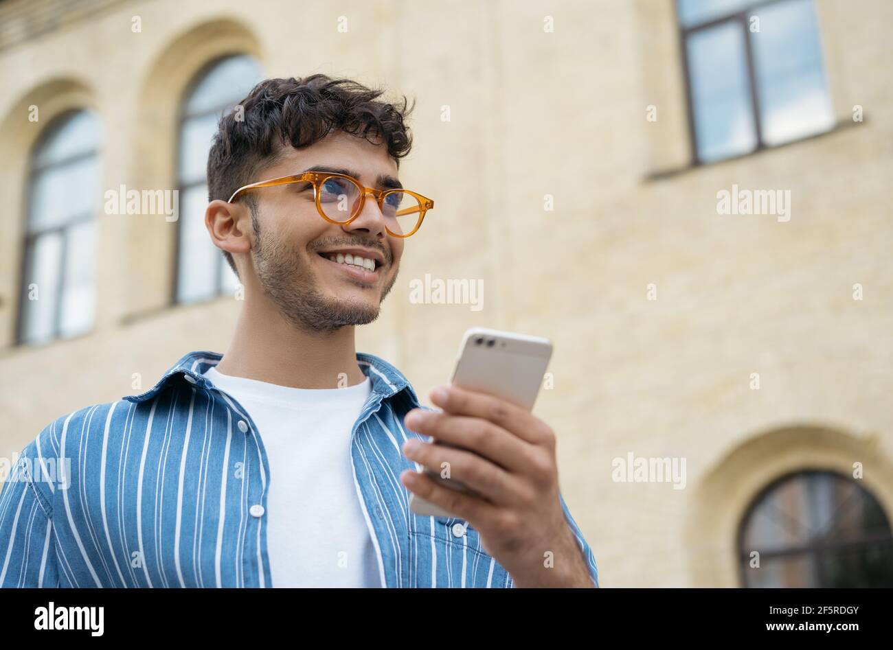 Indischer Mann, der mit dem Handy telefoniert, auf der Straße läuft und lächelt. Portrait von stilvollen Kerl tragen lässige Kleidung und Brillen mit Smartphone Stockfoto