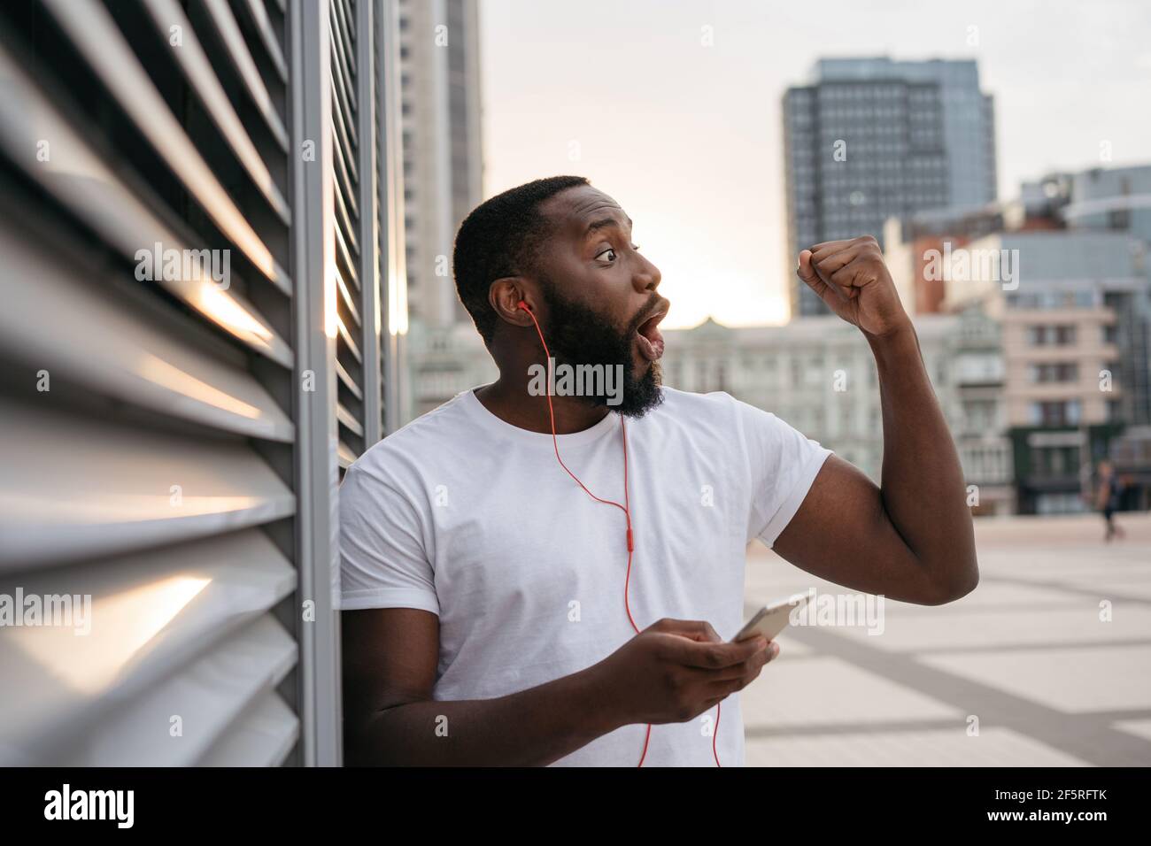 Happy Hipster Kerl trägt lässige Kleidung tanzen auf der städtischen Straße. Emotionaler afroamerikanischer Mann hält Handy und hört Musik im Freien Stockfoto