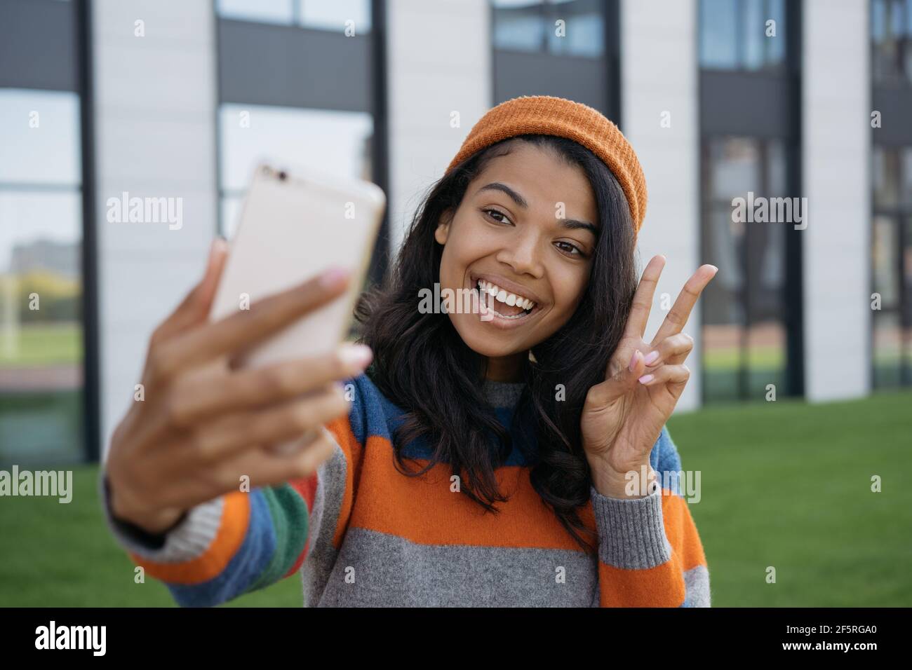 Junge emotionale Blogger Influencer mit Handy, Streaming-Video online. Lächelnde afroamerikanische Frau, die Selfie macht und ein Siegeszeichen zeigt Stockfoto