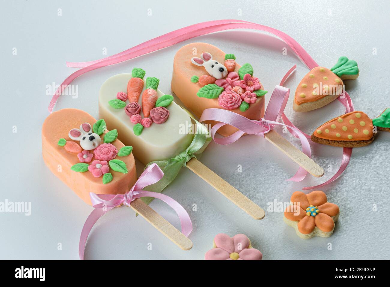 Karottenkuchencakesicles, verziert mit Kaninchen, Blumen und Schleife. Neben Karotte und blütenförmigen Cookies auf weißem Hintergrund. Stockfoto