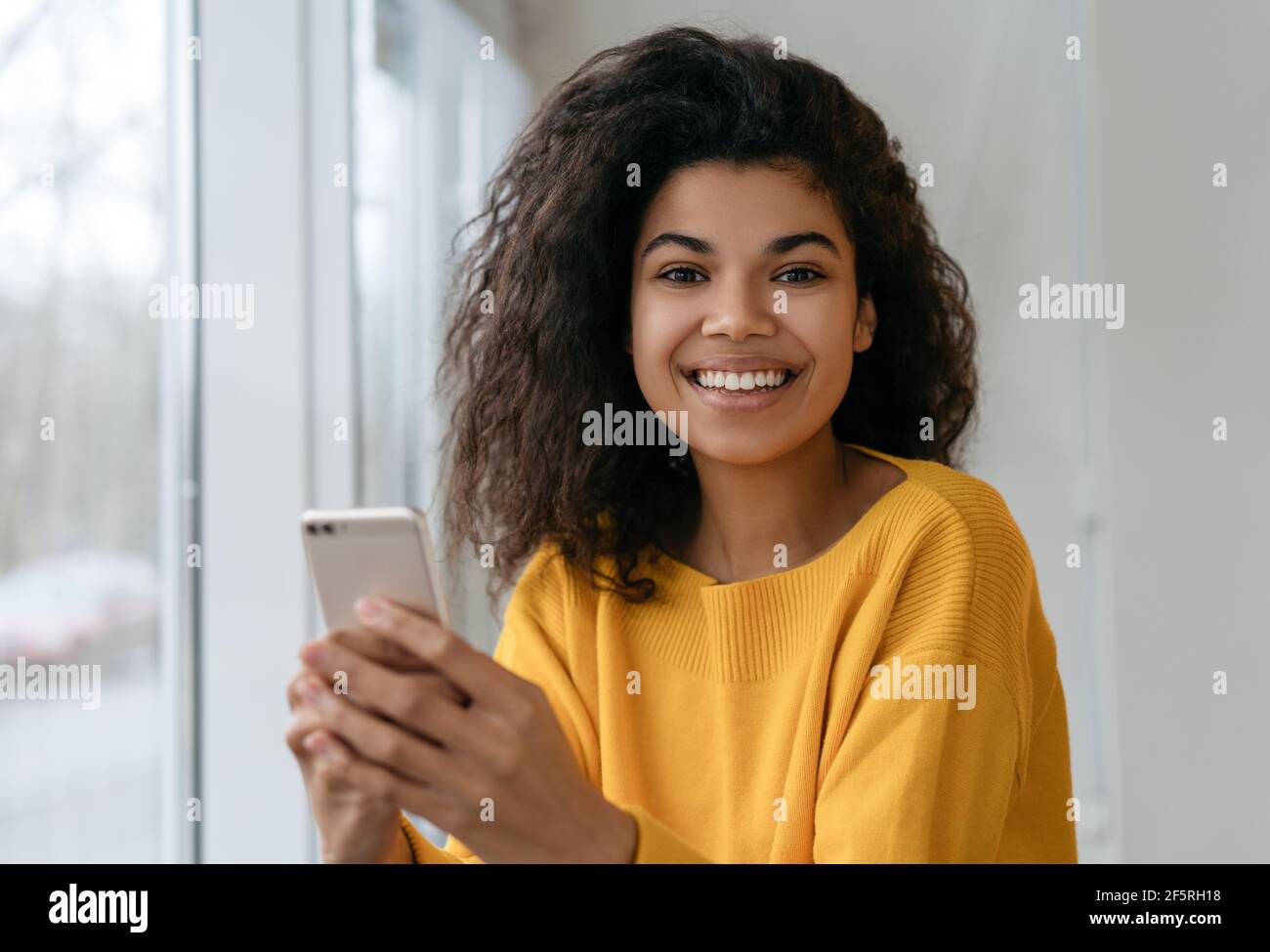 Schöne afroamerikanische Frau mit Handy, online einkaufen. Lächelnder Hipster hält Smartphone, bestellt Essen, blickt auf die Kamera Stockfoto