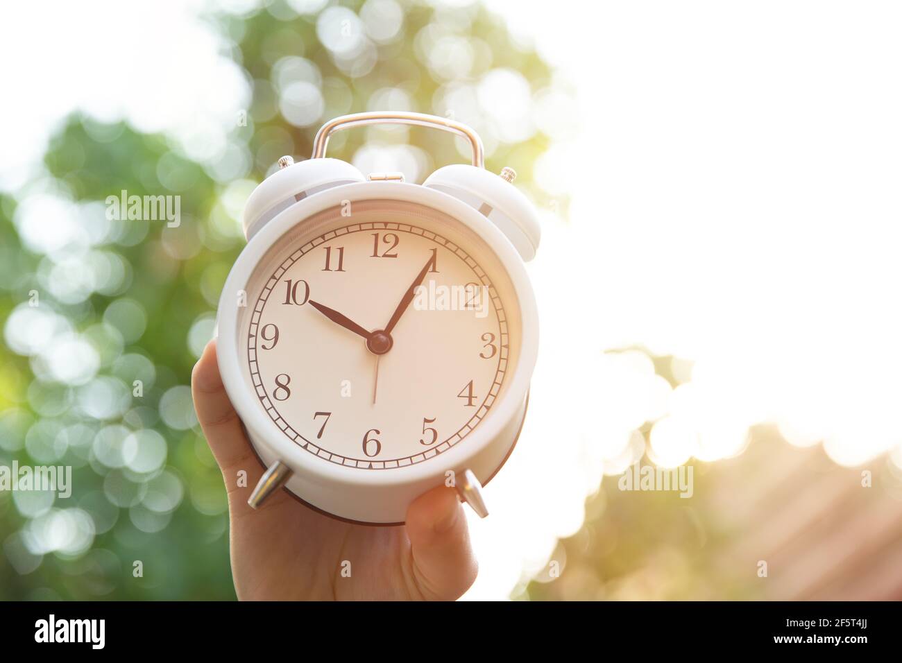 Morgenzeitkonzept, Hand halten weiße Glocke Uhr gegen Natur Sonne Licht Hintergrund. Stockfoto