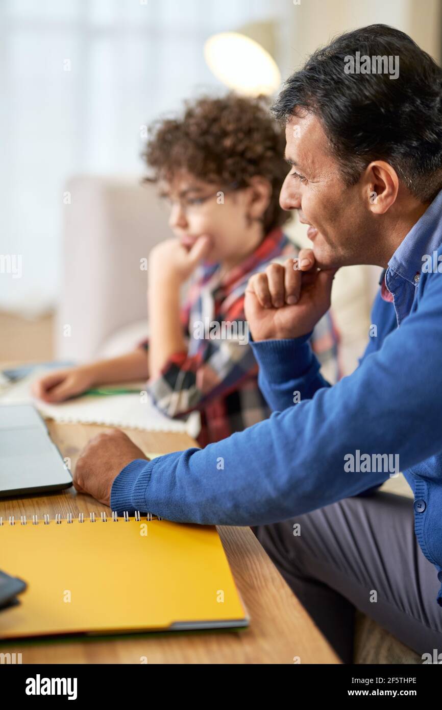 Das Kind auf den Erfolg vorbereiten. Lateinischer Vater mittleren Alters Überprüfung Hausaufgaben, hilft seinem niedlichen Sohn, Schuljunge mit Studien, während er am Tisch sitzen Stockfoto