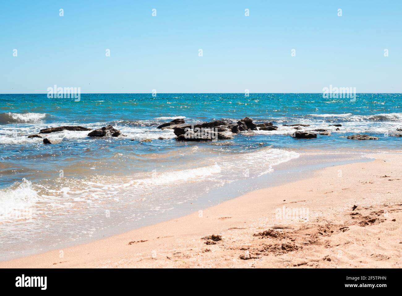 Meeresküste mit blauem Wasser gegen Steine und einem Sandstrand. Meer sonnige Sommerlandschaft. Stockfoto