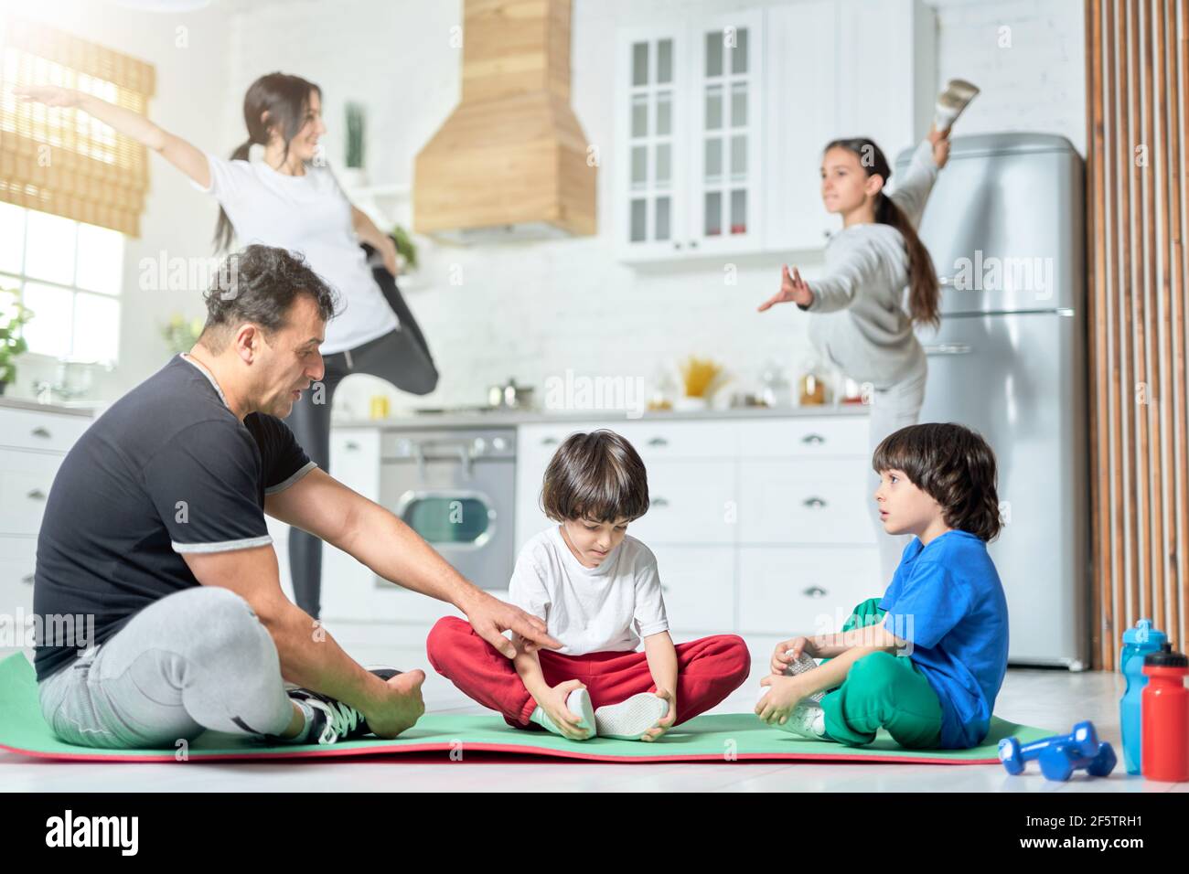 Körperlich fit. Zwei kleine Jungen sitzen auf einer Matte, üben Yoga mit Vater, während ihre Mutter und Schwester im Hintergrund trainieren Stockfoto
