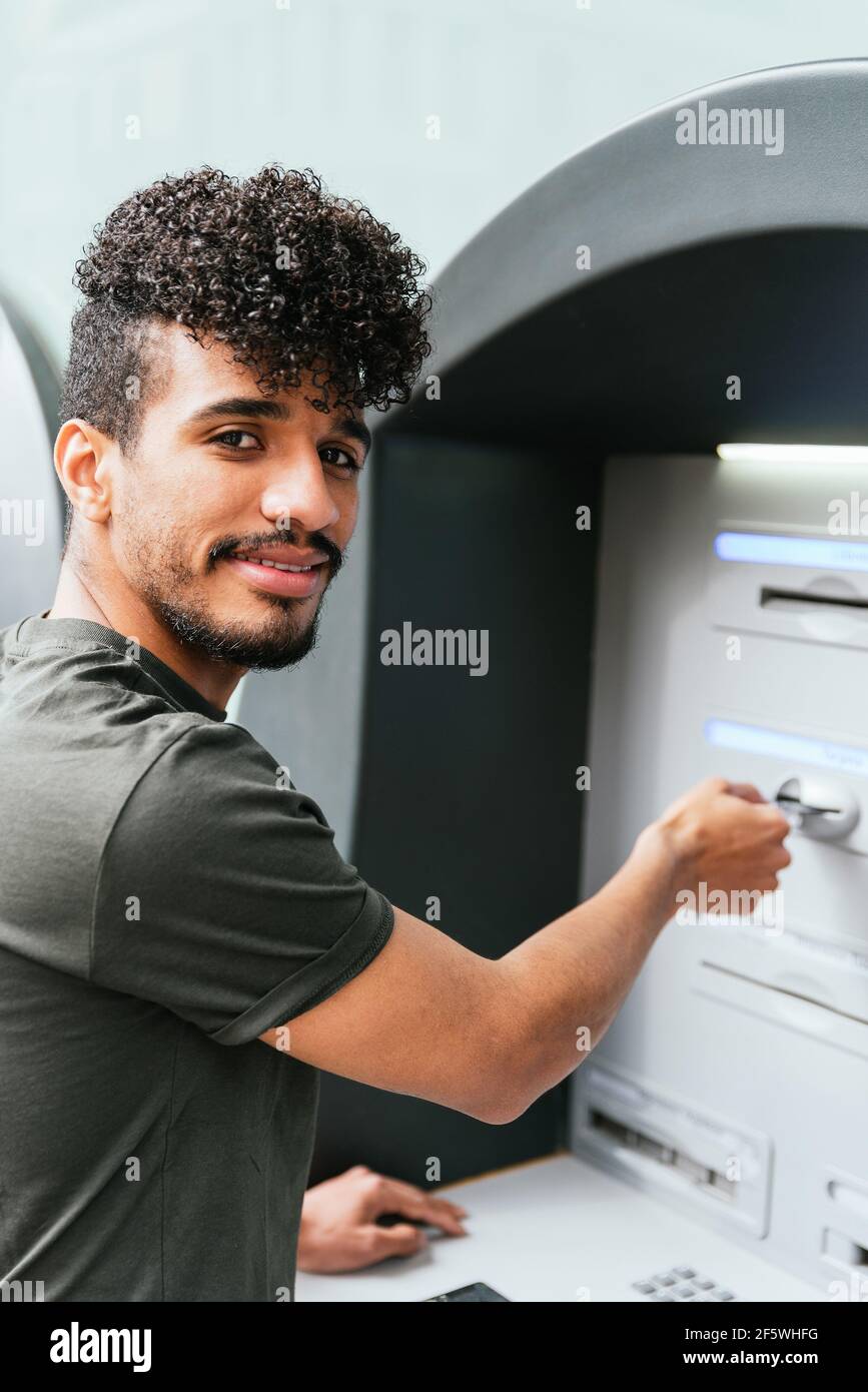 lateinamerikanischer junger Mann, der die Kamera anschaut und lächelt Mit einem Geldautomaten Stockfoto