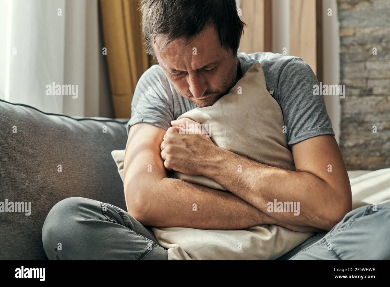 Deprimierter Mann, der auf dem Sofa im Wohnzimmer sitzt und sich umarmt, Porträt eines erwachsenen Mannes mit Angstanfall Stockfoto