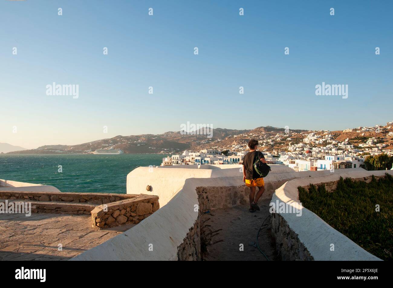 Ein junger Mann mit einem Rucksack auf der Schulter geht entlang einer malerischen Straße auf der Insel Mykonos in Griechenland. Im Hintergrund das Meer und das Dorf mit Stockfoto