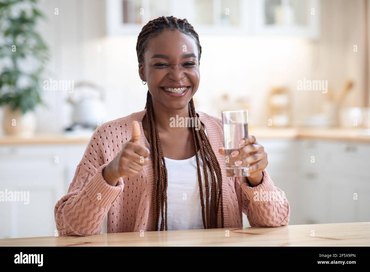 Fröhliche Schwarze Dame Trinkwasser Aus Glas In Der Küche Und Daumen Nach Oben Zeigen Stockfoto