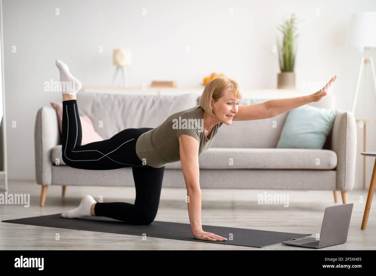 Heimsport. Eine ältere Frau muss vor dem Laptop arbeiten und ihr Bein und ihren Arm im Wohnzimmer anheben Stockfoto