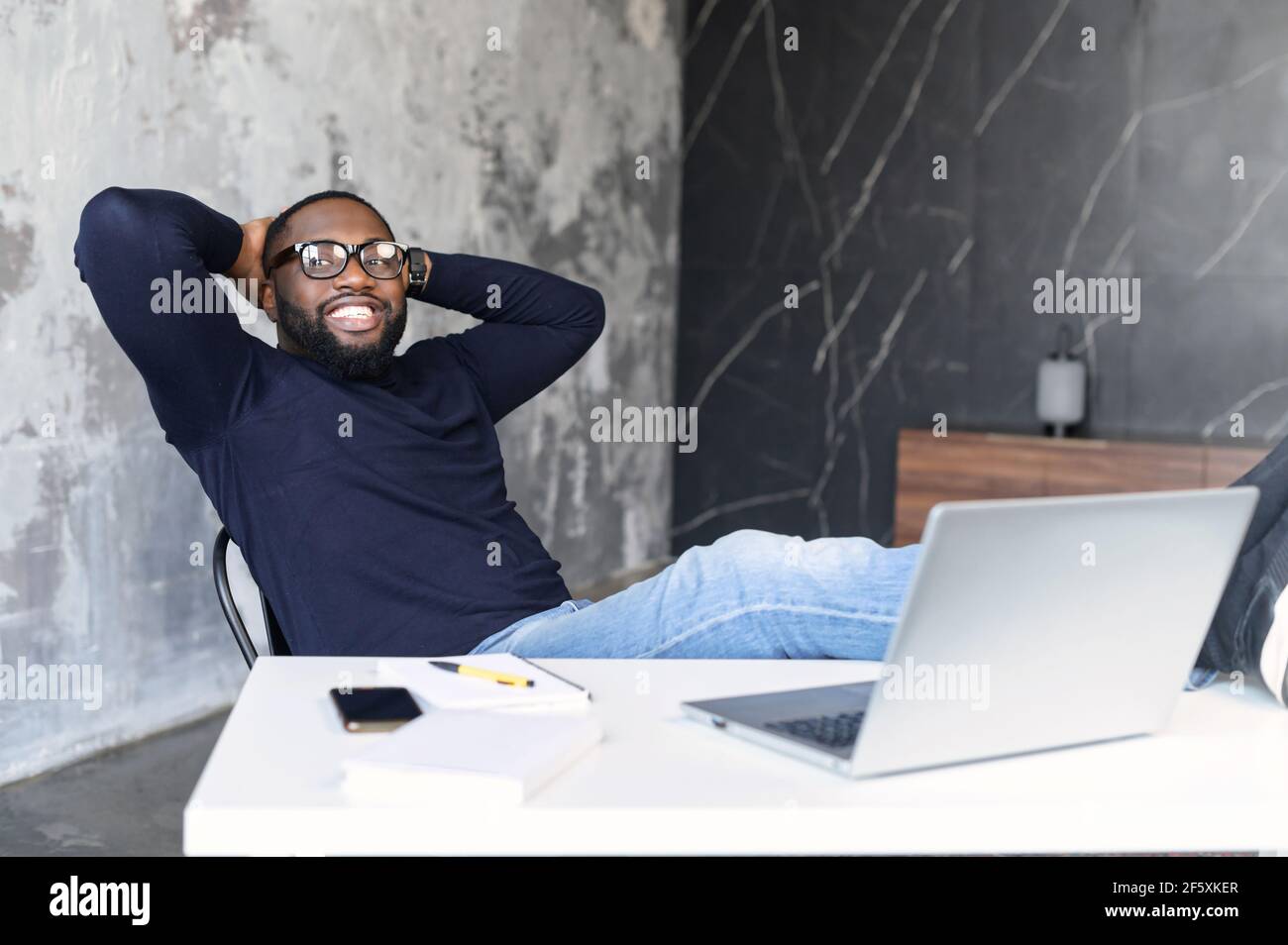 Glücklicher reifer afroamerikanischer Mann, der am Arbeitsplatz mit Laptop und Füßen auf dem Tisch sitzt und seine Hände hinter dem Kopf hält. Junge IT-Spezialisten machen eine Pause von der Arbeit. Entspanntes Arbeiten in geschlossenen Räumen Stockfoto