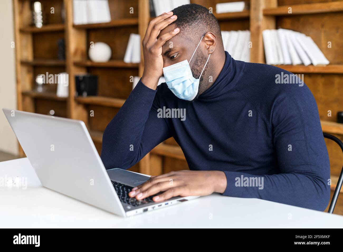 afroamerikanischer trauriger Geschäftsmann, der in medizinischer Maske am Laptop sitzt, bedeckt sein Gesicht mit der Hand, besorgt über finanzielle crysis, verlor sein Geld, fand heraus über Quarantäne und Geschäftsschließung Stockfoto