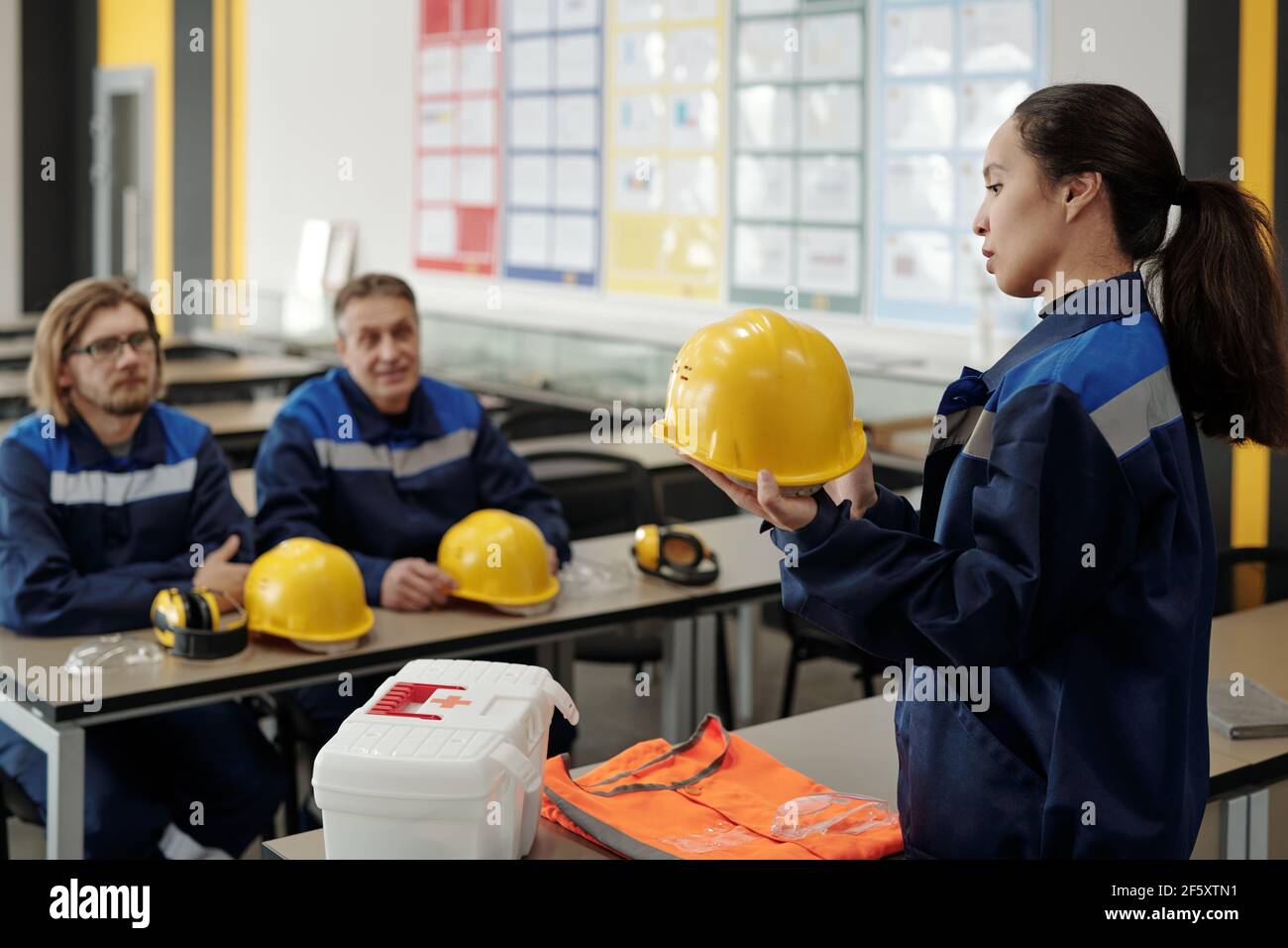 Junge hübsche Arbeiterin in Uniform, die vor dem Hotel steht Ihre männlichen Kollegen während der Beschreibung Zweck der Verwendung von Schutz Helm Stockfoto