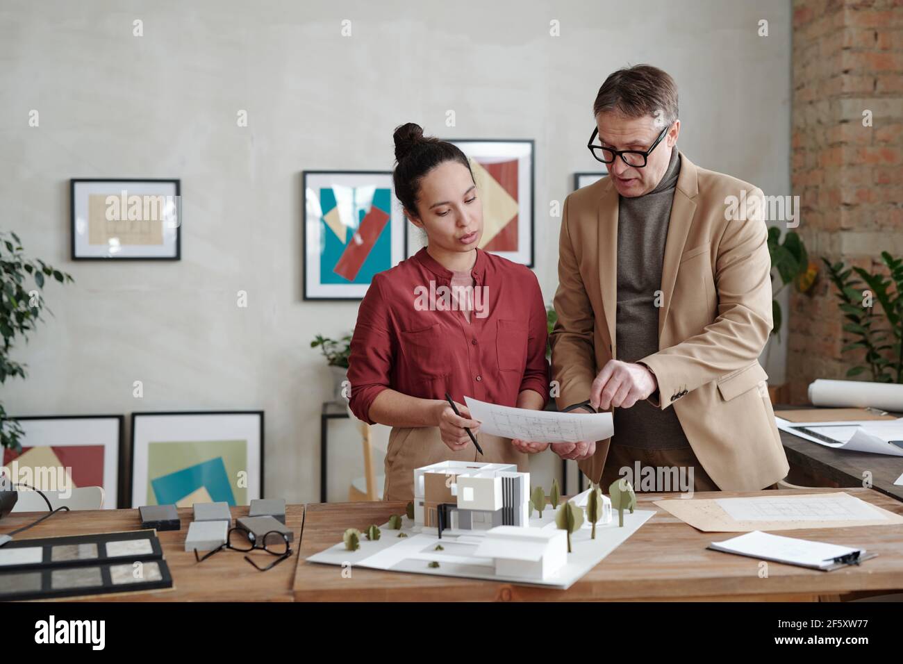 Junge Architektin und ihr reifer männlicher Kollege halten Papier Über dem Modell des neuen Hauses und des Hofes beim Besprechen der Details Der Skizze Stockfoto