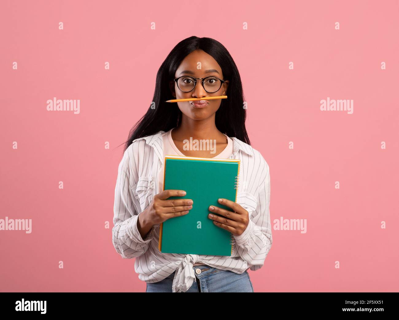 Dumme afroamerikanische Studentin mit Notizbüchern, die Bleistift dazwischen halten Ihre Lippen und Nase auf pinkem Studiohintergrund Stockfoto