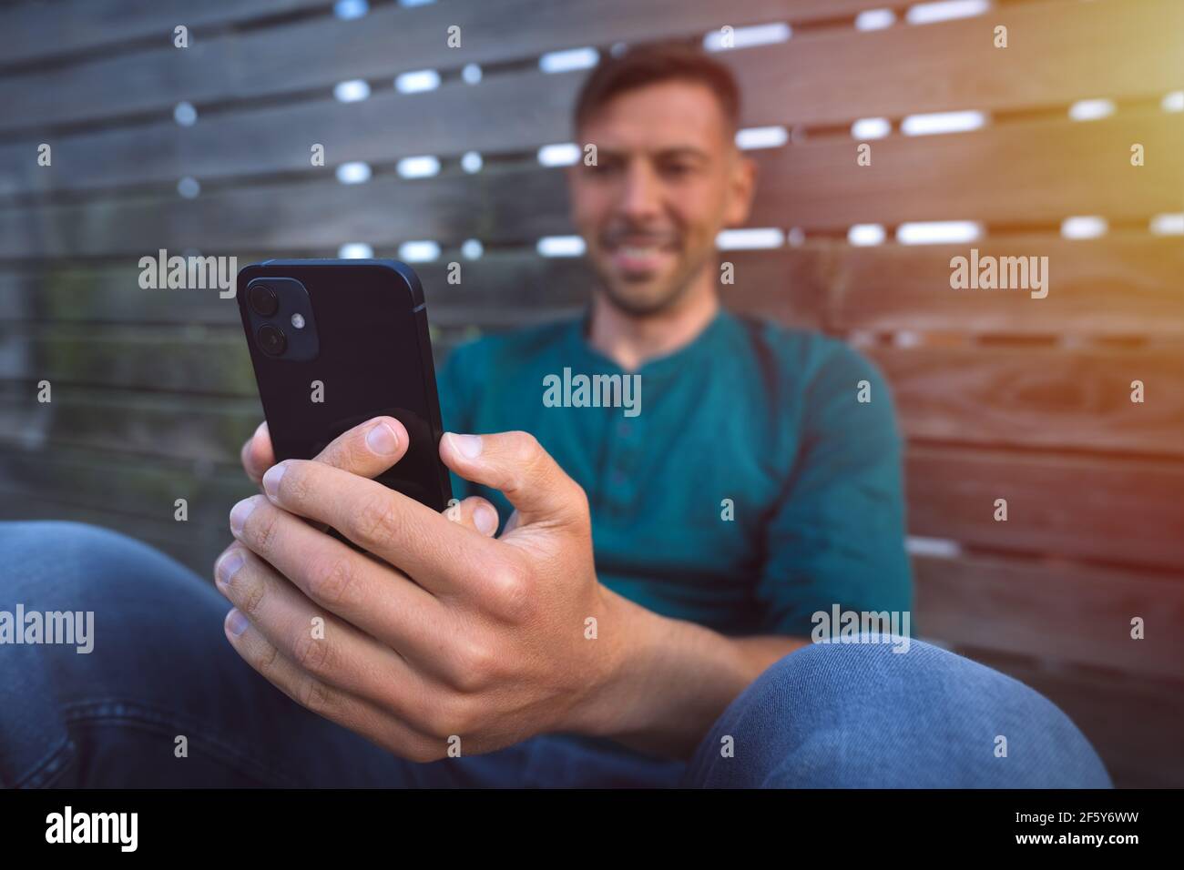 Junger lächelnder Mann mit seinem schwarzen Smartphone im Freien bei Sonnenuntergang. Smartphone betrachten. Mann mit einem facetime Videoanruf Stockfoto