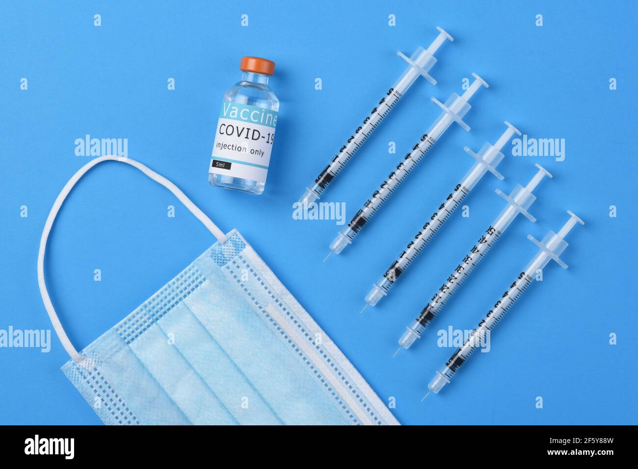Flacher Schuss von Spritzen ein Fläschchen mit Covid-19-Impfstoff und eine OP-Maske auf blauem Hintergrund. Stockfoto
