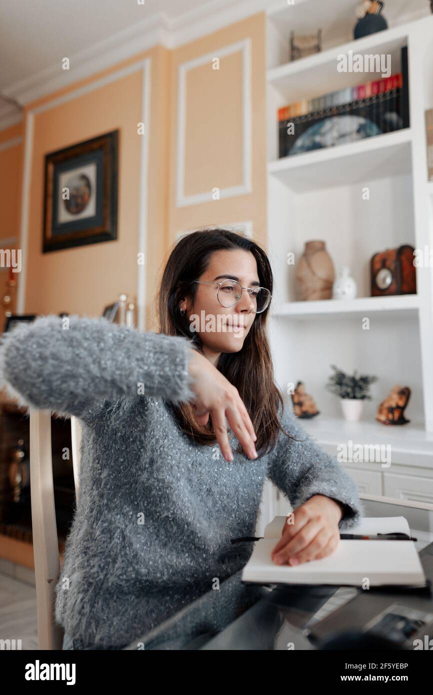 Medium shot junge taube Frau trägt eine Brille und ein Pullover ist mit Gebärdensprache Namen buchstabieren, während in einem Online-Videoanruf in ihrem Wohnzimmer. Stockfoto