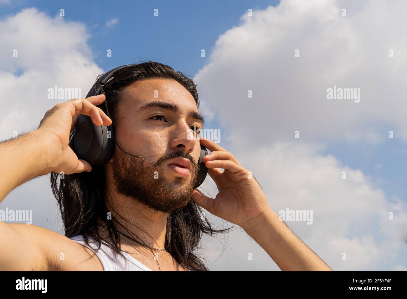 Bärtiger junger Mann, der kabellose Kopfhörer mit dem Himmel im Hintergrund trägt, über das Leben nachdenkt, meditiert Stockfoto