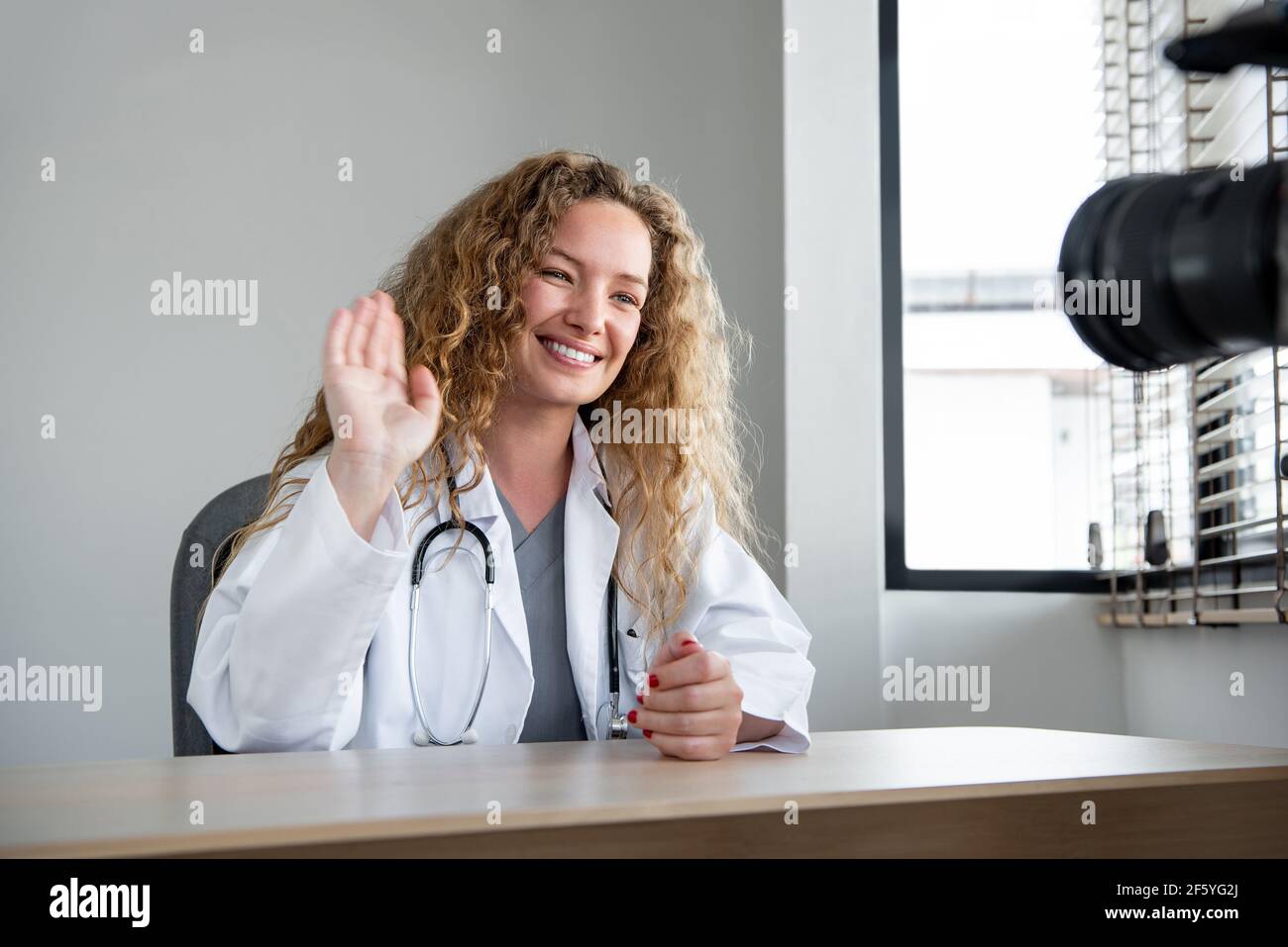 Lächelnd fröhlich kaukasischen Frau Arzt winkt Hand zur Kamera, während Live-Streaming aus dem Krankenhaus Stockfoto