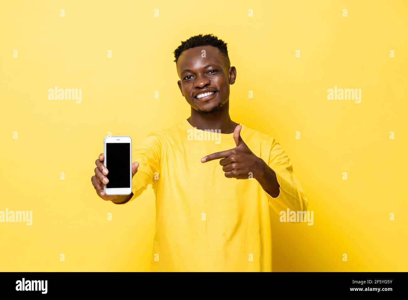Lächelnder junger afrikanischer Mann, der die Hand auf das Mobiltelefon zeigt Gelber isolierter Studiohintergrund Stockfoto