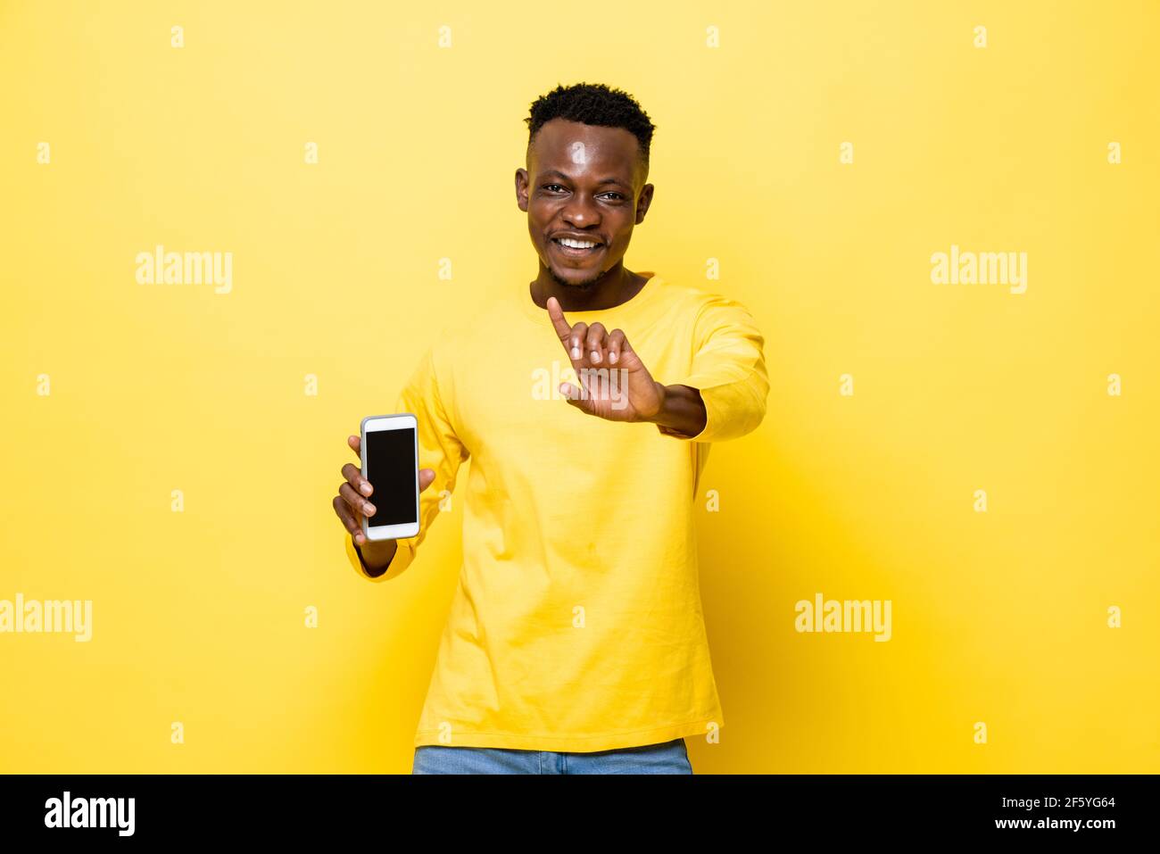 Junger lächelnder afrikanischer Mann hält Handy und tut Nein Geste in gelb isolierten Studio-Hintergrund Stockfoto