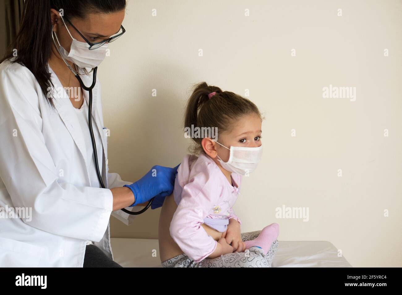 Mollige kleine Mädchen in der pädiatrischen Untersuchung durch ihren Arzt. Der Arzt hört auf Ihre Lungen und Herz. Sie tragen schützende Gesichtsmasken Stockfoto