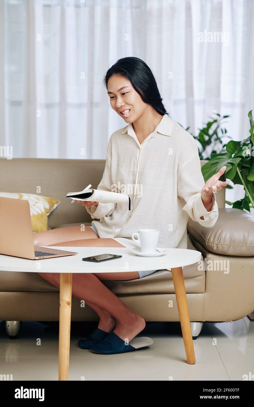 Ziemlich junge asiatische weiblich Student im Pyjama sittingon Sofa an Startseite und Beantwortung der Frage des Lehrers, wenn mit Online-Klasse Stockfoto
