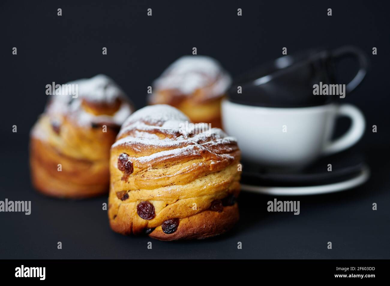 Frische Osterbäckerei oder Tee Zeit Konzept. Süße leckere Cruffins mit Rosinen und weißem Zuckerpulver darauf mit schwarzen und weißen Kaffeetassen in ihrer Nähe. Schwarzer Hintergrund. Hochwertige Fotos Stockfoto