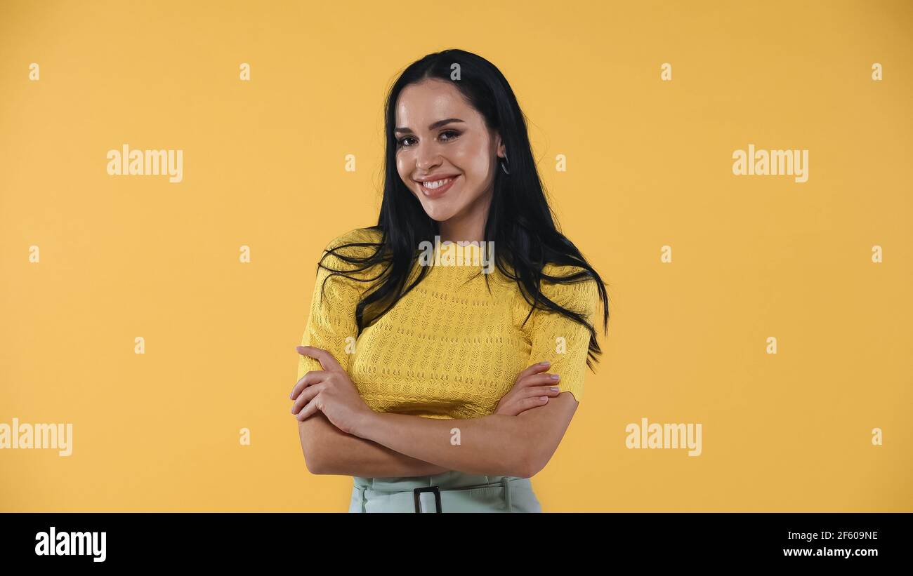 Flirty, fröhliche Frau mit gekreuzten Armen stehend, während lächelnd auf Kamera isoliert auf gelb Stockfoto