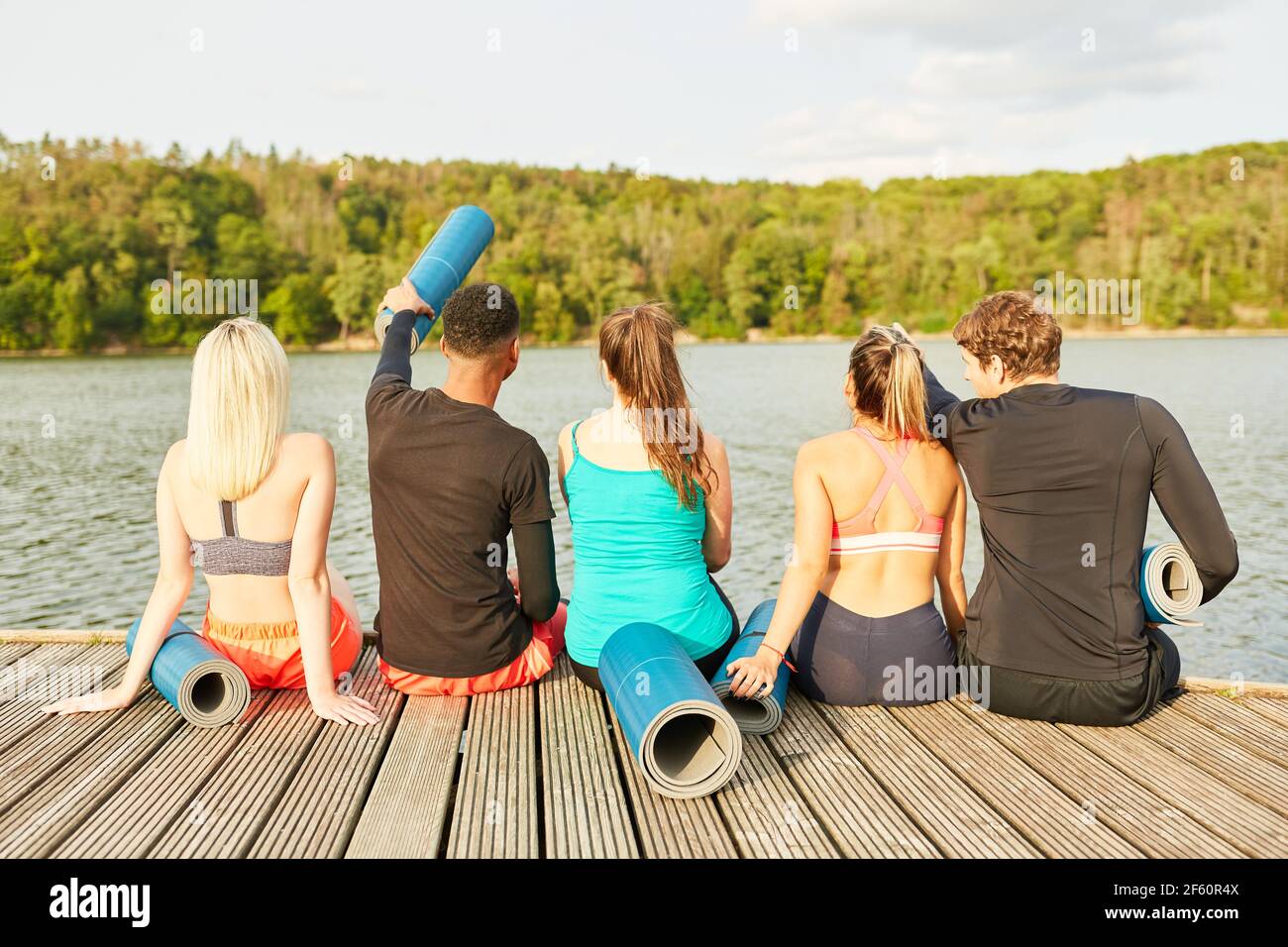 Gruppe von jungen Menschen mit Yoga-Matten sitzen auf einem Holzsteg am Seeufer in der Natur Stockfoto