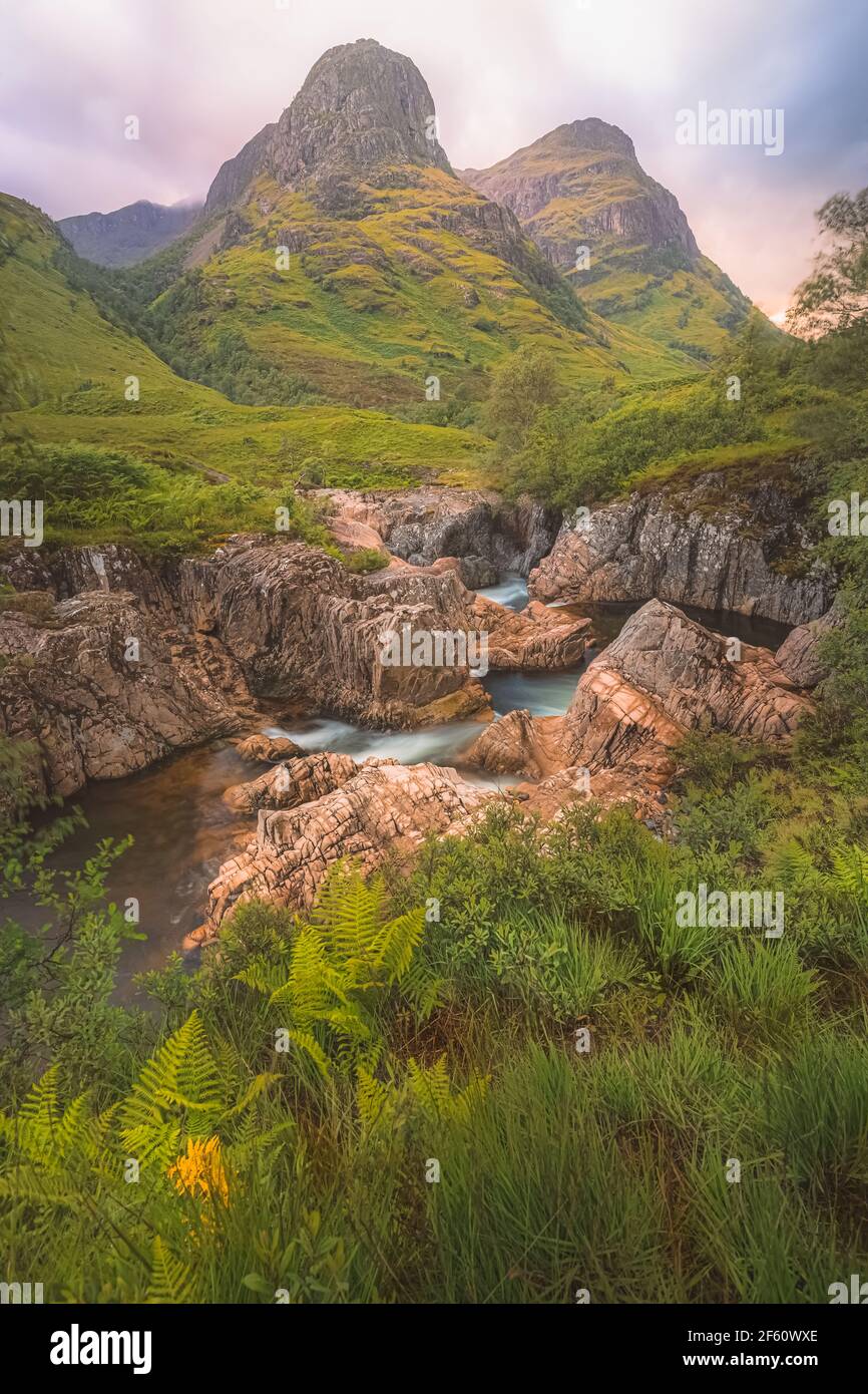 Idyllische Berglandschaft entlang des Flusses Coe zu den Three Sisters of Glencoe mit einem bunten Sonnenuntergang oder Sonnenaufgang in den schottischen Highlands, Schottland. Stockfoto